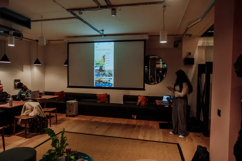 A woman stands near a projector screen showing a phone app demo in the event space.