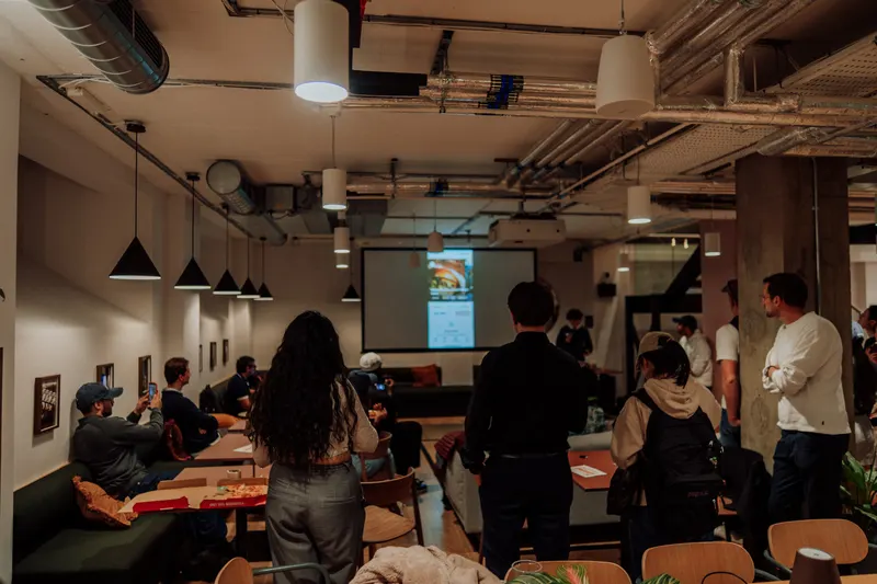 Guests stand and sit at the back of the room watching a founder present on screen.