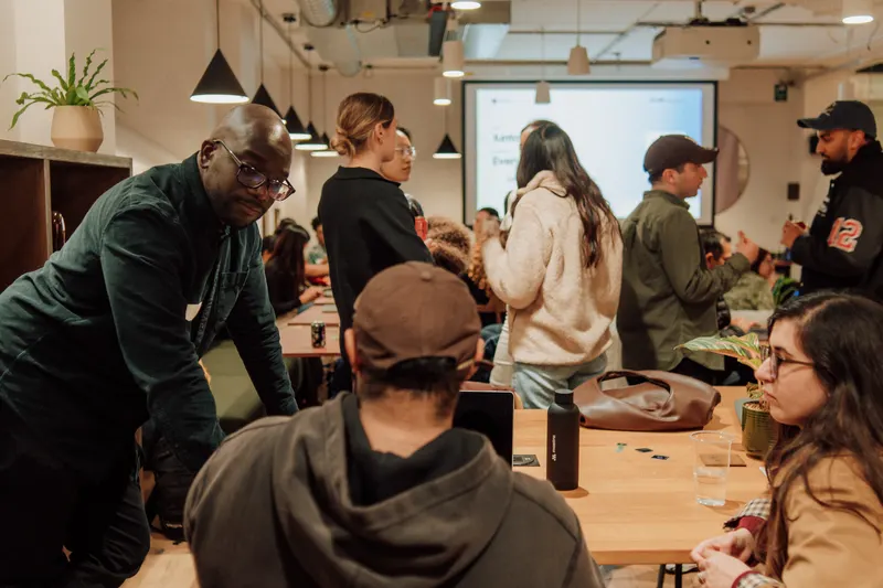 A man in a dark green jumper leans over a table of founders as the room fills.