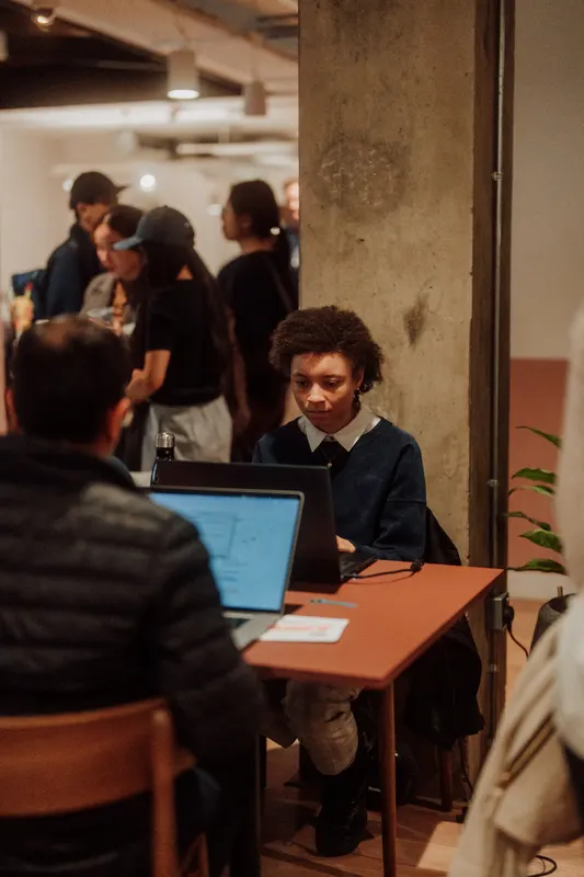 A founder works on a laptop at a small desk while other guests stand behind.