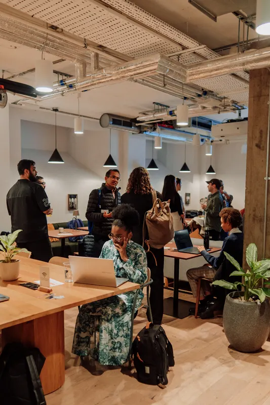 Founders stand and chat among tables and plants while one person works on a laptop.