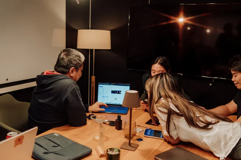 Founders huddle around a laptop at a wooden table in a darker side room.