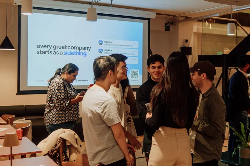 Founders chat in front of a projected slide reading every great company starts as a sidething.