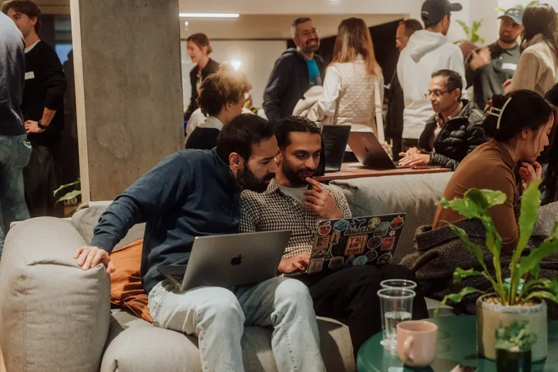 Two founders share a laptop on a grey sofa with guests chatting in the background.