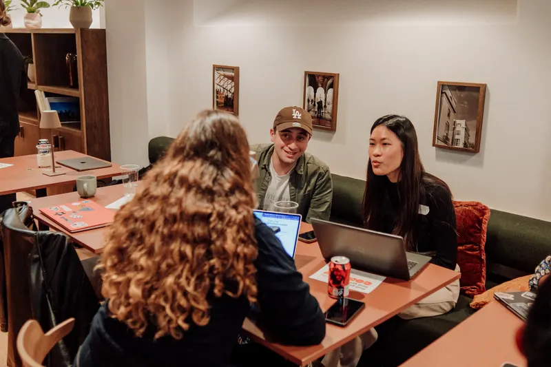 Two founders smile at a woman with curly hair across a table with laptops and drinks.