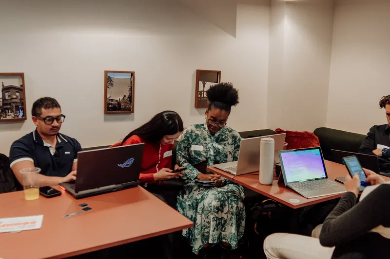 Founders sit at a table with laptops and phones, chatting and comparing their side projects.