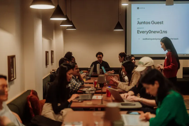 Founders sit around a long table with laptops while a speaker stands by the screen.