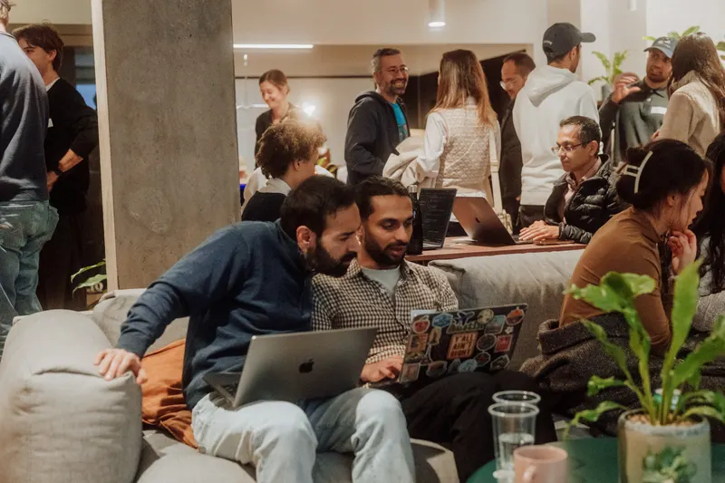 Two founders look at a laptop covered in stickers on a sofa while others mingle behind.