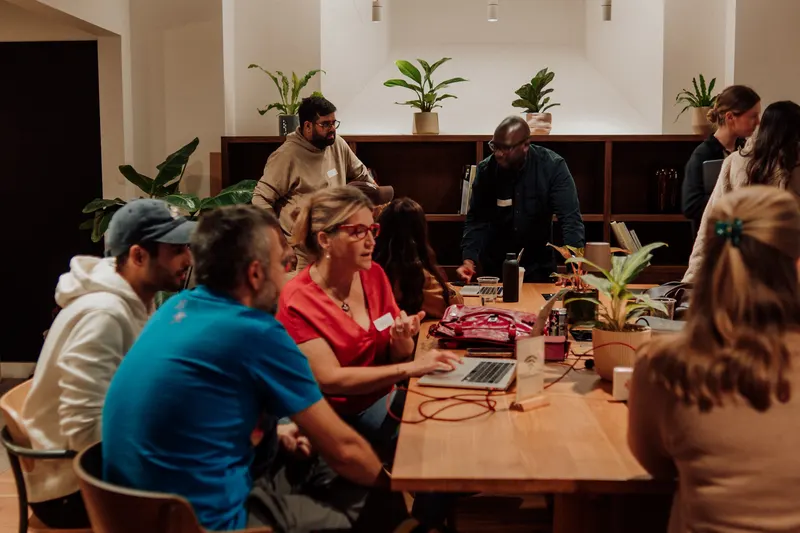 Founders sit around a table with a laptop and cables while others walk past behind them.