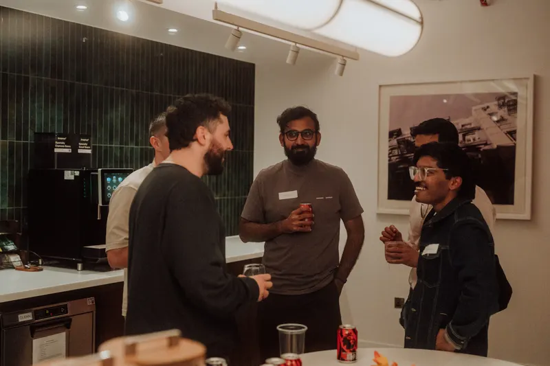 Three founders stand talking by the coffee area, one holding a drink and smiling.