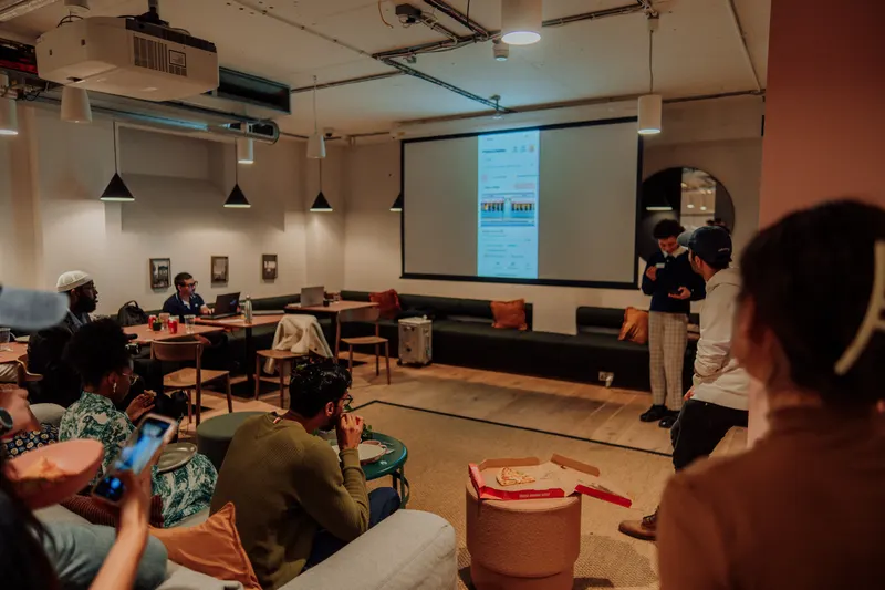 Founders sit with pizza boxes and phones watching a presenter beside the projected app demo.