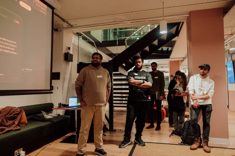 Four founders stand at the back of the room watching a presentation beside the staircase.