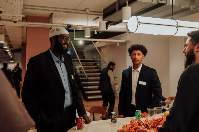 Two guests in blazers chat by a table with cans of drink and autumn leaves.