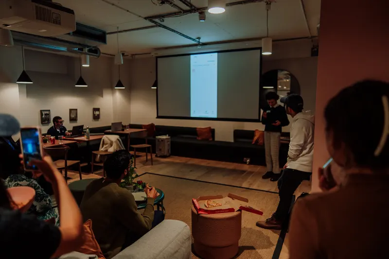 Guests film a founder demoing an app on screen while pizza boxes sit on the table.