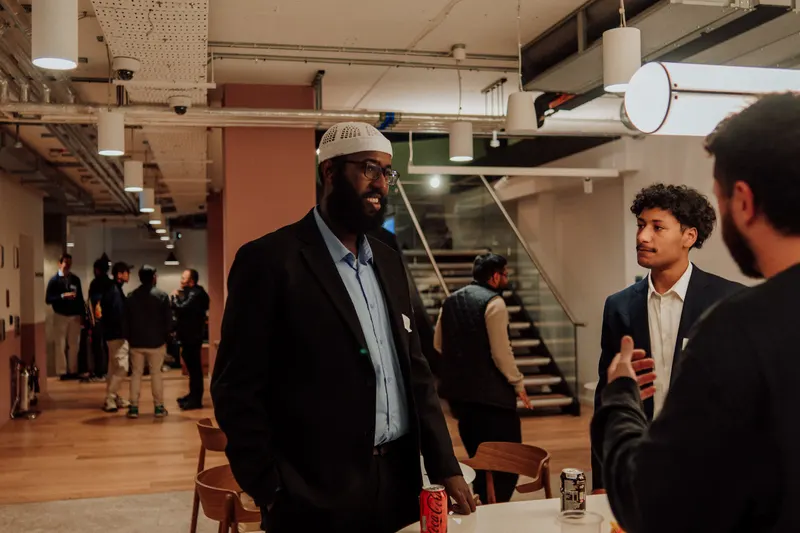 A bearded man in a white cap talks with a younger man in a blazer near the stairs.