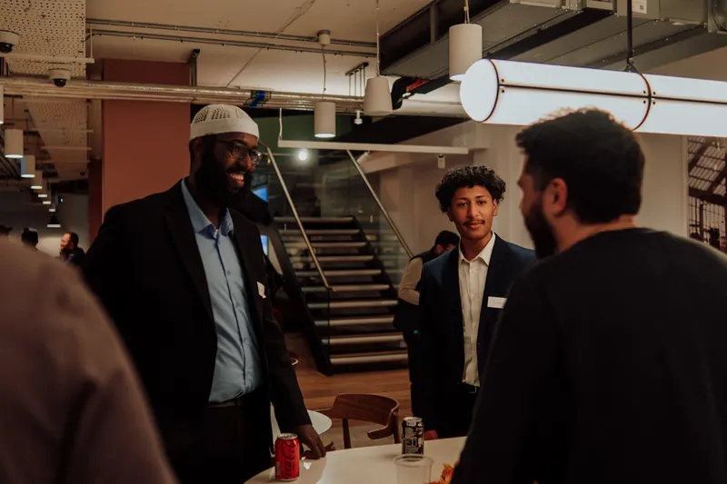A man in a white cap and black blazer smiles while talking to guests by the stairs.
