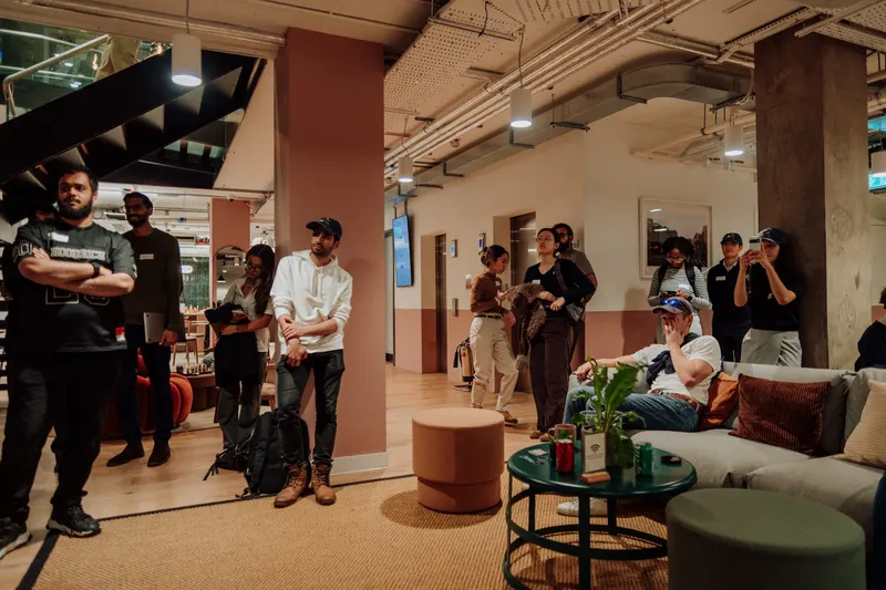 Guests stand near the staircase watching a talk while others sit on a sofa.