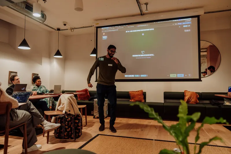 The host stands with a microphone and gestures as guests sit on sofas behind a plant.