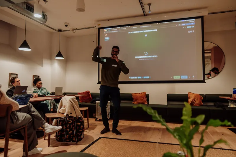 The host raises a hand while speaking into a microphone beside the large projector screen.