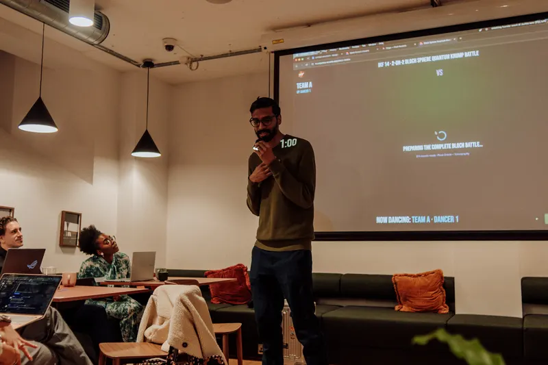A man holds a microphone and speaks beside a projected screen while guests watch from sofas.