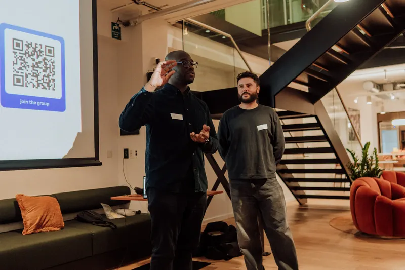 One host gestures while speaking, standing beside another, next to a projected QR code slide.