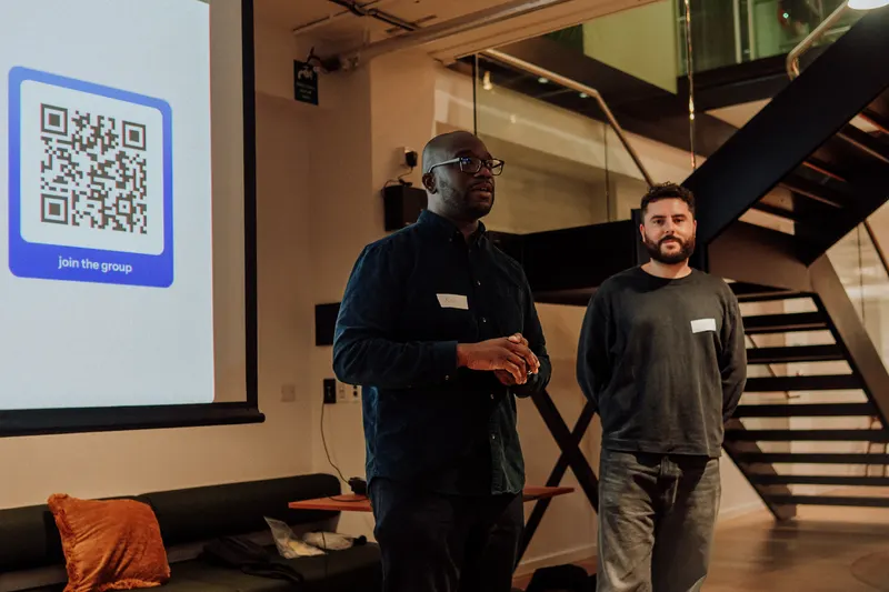Two hosts stand by the staircase next to a slide with a QR code to join the group.