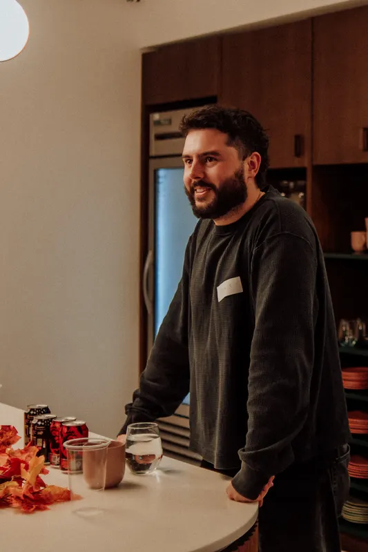 A bearded man in a dark jumper leans on a high table and smiles at someone off camera.