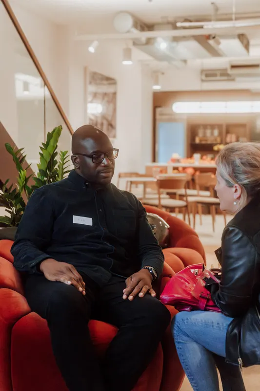A man in glasses sits on a red armchair and talks with another guest.
