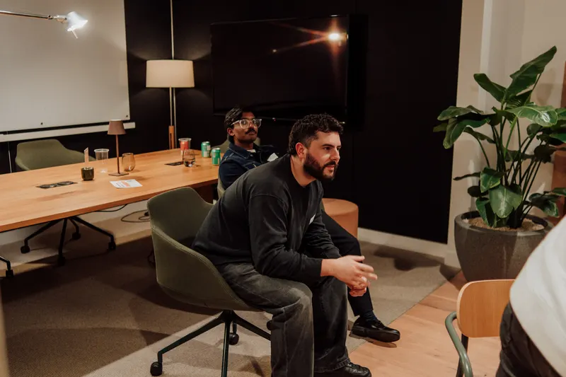 A bearded man sits forward on a chair, watching a presentation with a tall plant beside him.