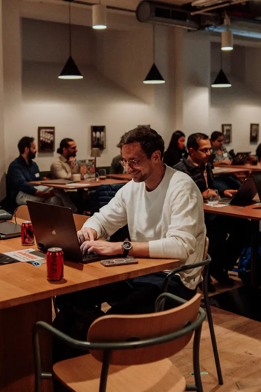 A man in a white jumper smiles while working on a laptop at a table.