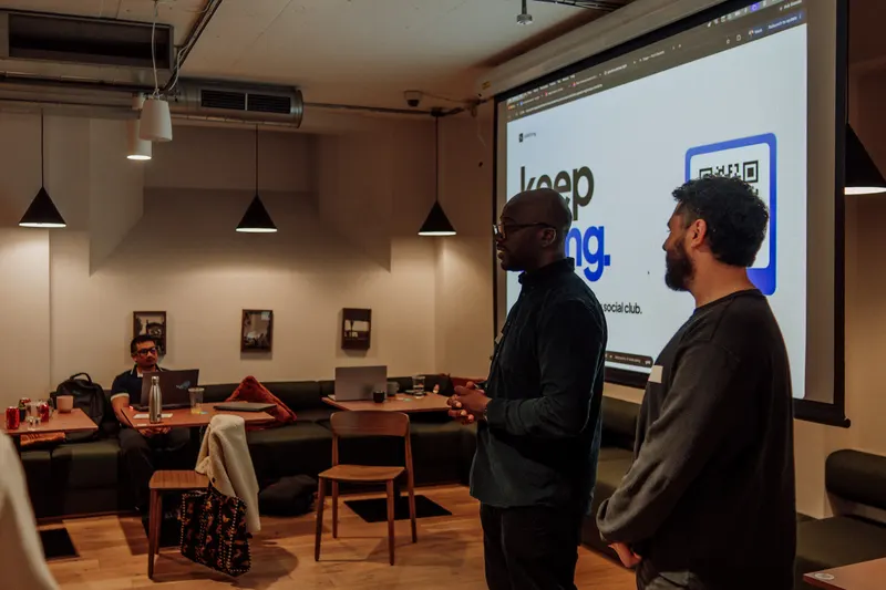 Two men stand beside a large screen showing a keep going slide while others sit behind.