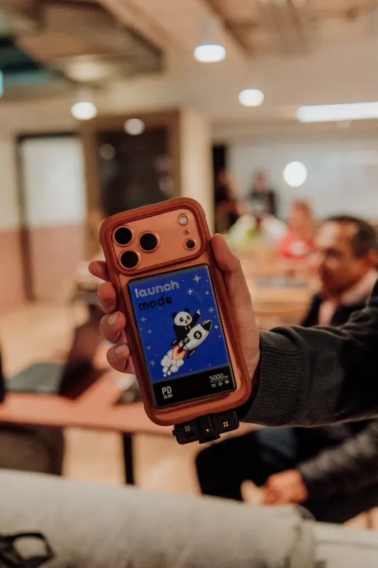 A phone in an orange case shows a launch app screen while guests chat behind it.