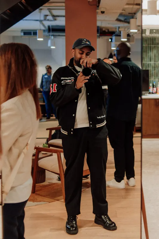 A man in a bomber jacket takes a photo with a camera while guests chat nearby.
