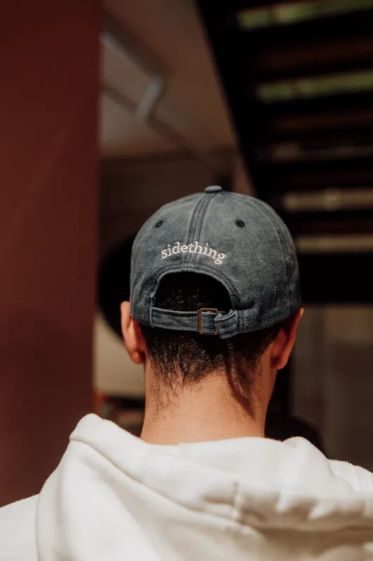 The back of a faded sidething cap worn by a man in a white hoodie.