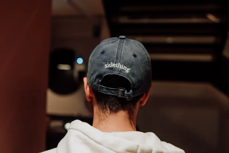 A man in a faded cap marked sidething and a white hoodie seen from behind.