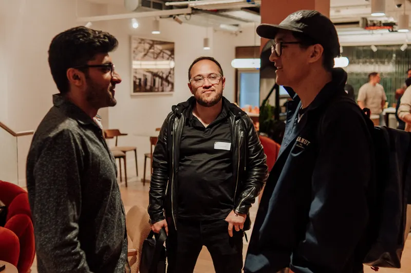 Three men in dark jackets stand close together chatting at Growth Night in London.