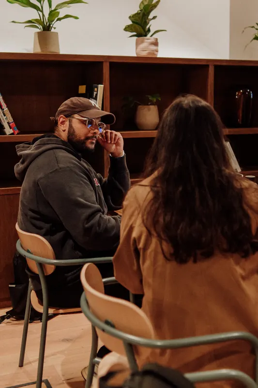 A man in a cap and a woman with long hair talk near a bookshelf.